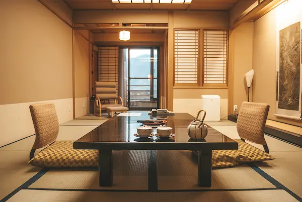 日本の温泉旅館の部屋の写真。ローテーブルが置かれ、ライトは暖色で、座椅子が三つ並んでいる。テーブルの上には急須と湯呑みが置かれている